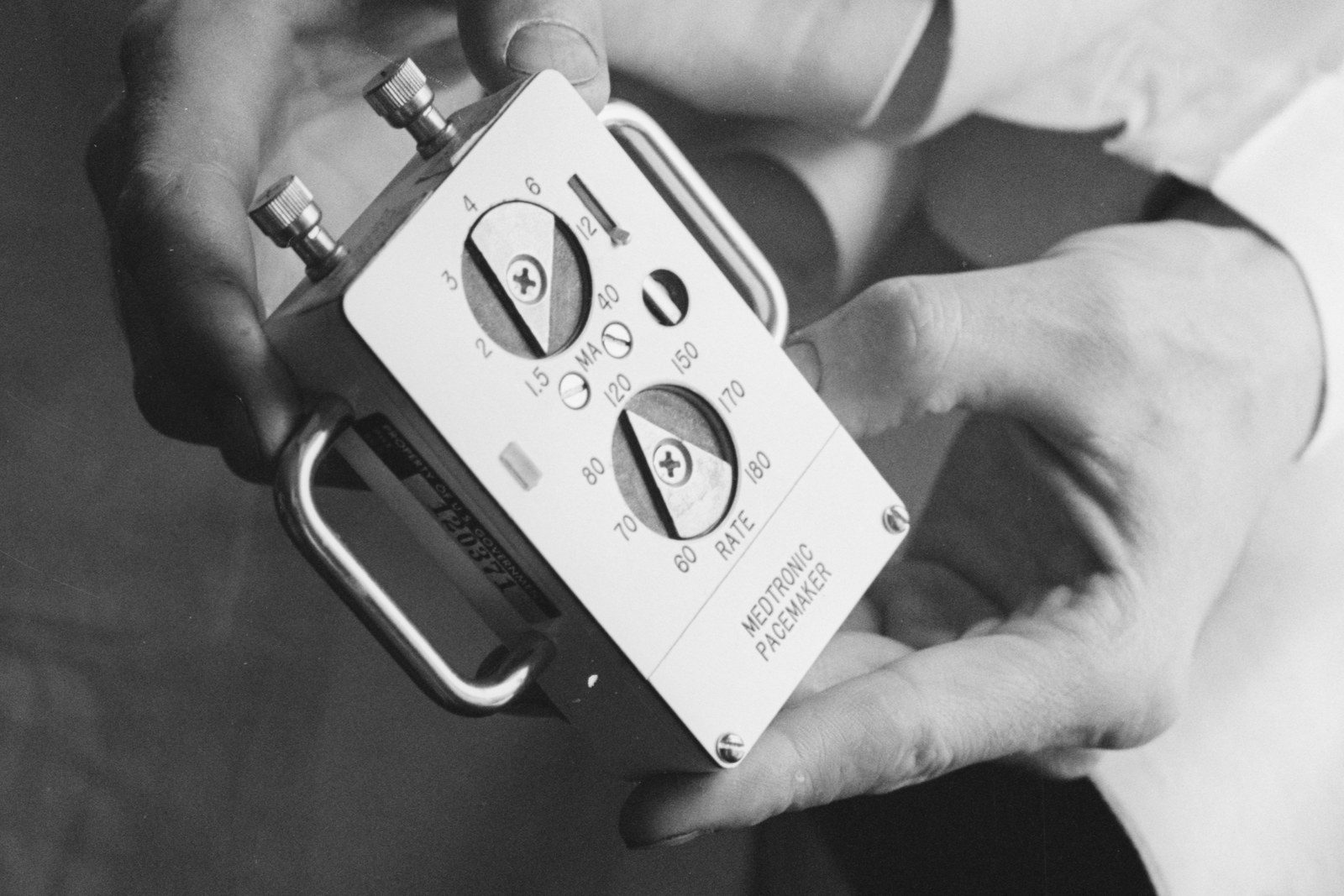 A doctor holds an early Medtronic pacemaker in 1961, its RATE and milliamp dials visible on the faceplate.